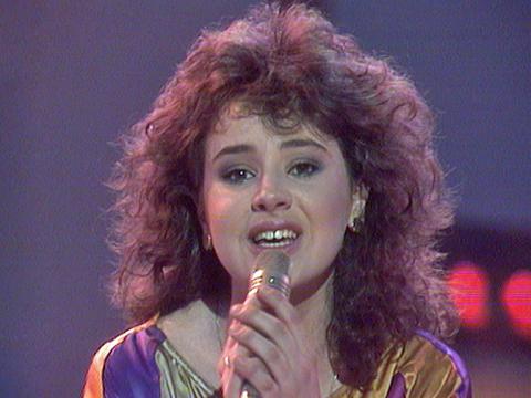 A head and shoulders shot of a teenage girl holding a microphone and singing, looking directly at the camera. 