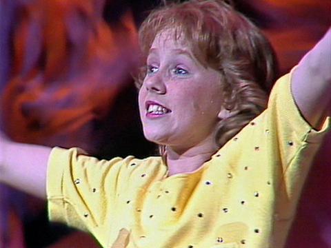A young teen girl with strawberry blonde hair, wearing a yellow t-shirt is standing with her arms outstretched. 