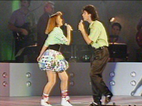 Full length image of a teenage girl and boy on stage facing each other. Both are singing into microphones.