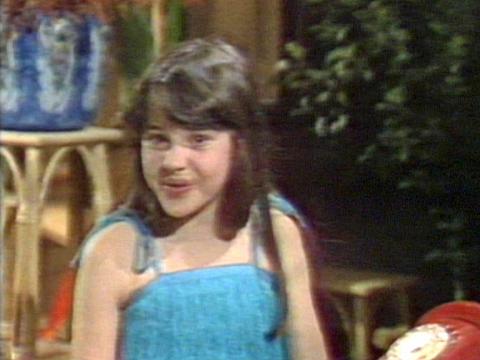 A young girl in a blue top is singing and looking at the camera. She has a red rotary telephone in front of her.