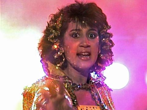 A head and shoulders shot of a teenage girl wearing a gold outfit with bright pink backlighting. She is looking directly towards the camera. 