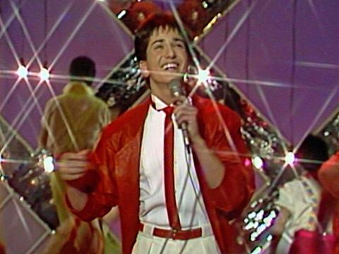 A teenage boy wearing a red jacket and red tie is smiling and singing into a microphone. He is on a stage with backup dancers behind him.