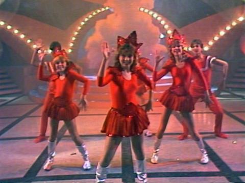 A group of six teenagers on stage dressed in red costumes performing a song and dance.