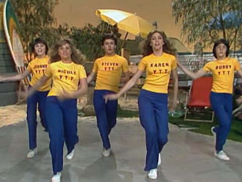 A group of teenagers on stage dancing. They are all wearing blue trousers, white sneakers and bright yellow T-shirts with their names printed on them.