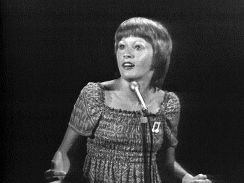 A teenage girl in a shirred dress stands at a microphone singing and looking slightly to her right.