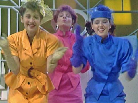 Three teenage girls in colourful 80s outfits doing a dance routine to camera