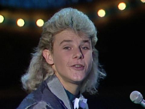 A teenage boy with blond hair looking at the camera while he sings into a microphone.
