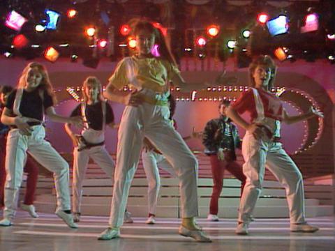 A group of teenagers on stage dancing in white trousers with colourful t-shirts and suspenders.