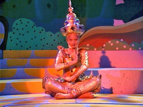 A young girl sitting cross-legged on a stage dressed in a traditional Thai costume holding a dance pose.