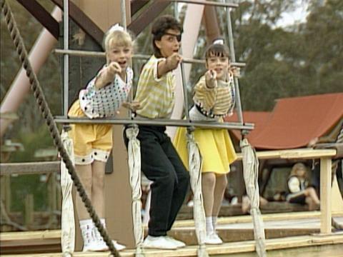 Three children at a theme park pointing towards the camera