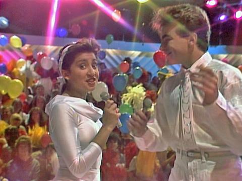 A teenage boy and girl dressed in white, holding microphones and facing each other. In the background there is an audience and lots of colourful balloons.