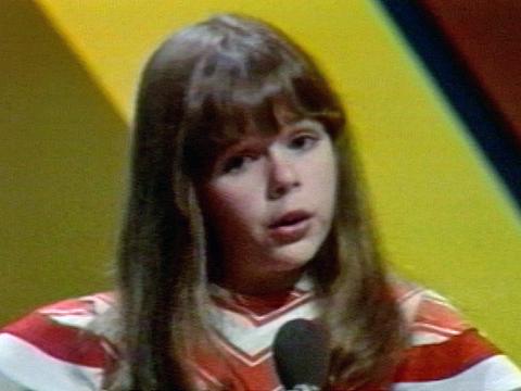 Head and shoulders shot of a young girl standing in front of a microphone. Her head is slightly tilted to her right and she looks like she is singing