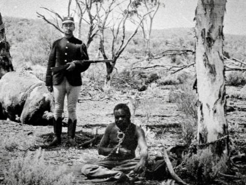 A soldier with a gun stands over an Aboriginal man on the ground in chains