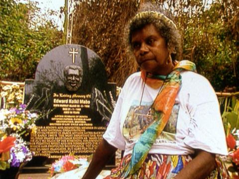 Bonita Mabo sits by the grave of her husband Eddie Mabo on Mer. She wears a bright coloured scarf and a t-shirt that reads 'I'm proud of you darling'.