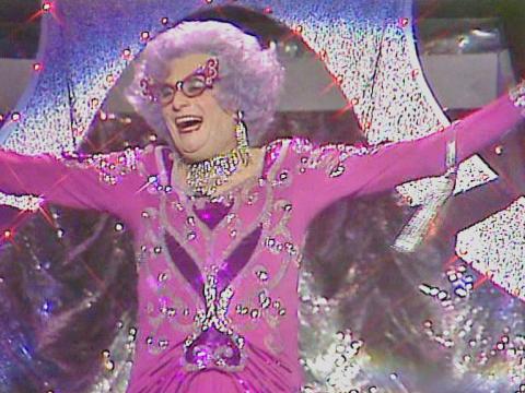Dame Edna Everage blowing kisses to the audience. She is wearing a spectacular pink frock.