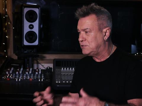 Singer Jimmy Barnes pictured from the chest up, in a black t-shirt and sitting in front of a mixing deck in a recording studio. 