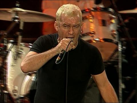 Jimmy Barnes wearing black t-shirt on stage, singing into a microphone. He is pictured from the waist up with a drum kit in the background.