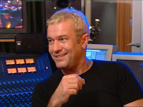 Head and shoulders shot of Jimmy Barnes wearing a black t-shirt, sitting in a studio in front of a mixing desk. 