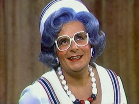 Comedian Barry Humphries dressed as his character Dame Edna Everage in a white blouse with red and blue stripes around the collar, matching pillbox hat, white-rimmed glasses and beaded necklace.