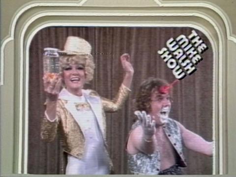 Jeanne Little is dressed in a golden tuxedo. A man is next to her with his arms outstretched and a grin on his face.