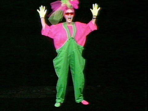 Jeanne Little is wearing a flouresent pink and green outfit. She has her hands up in the air.