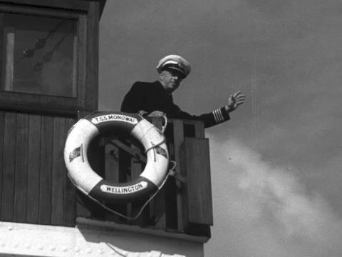 A sea captain waves from a ship's deck. The life preserver says TSS Monowai.