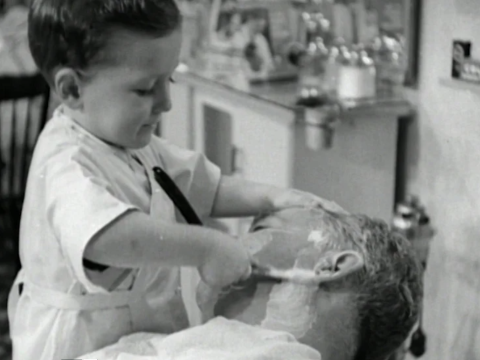 A small boy in a barber's apron is giving a man a shave with a razor.