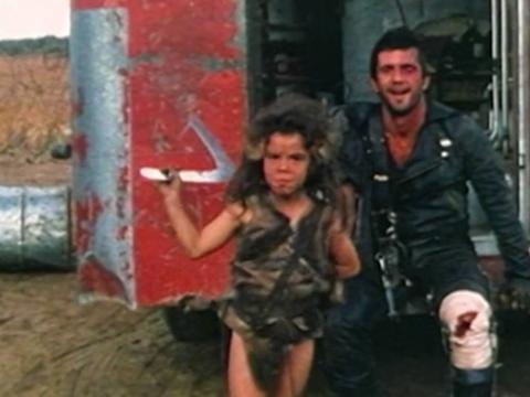 Actors Emil Minty and Mel Gibson in costume on the set of Mad Max 2. They are seated on the back of trailer in between takes and Minty is holding a silver boomerang while Gibson is laughing
