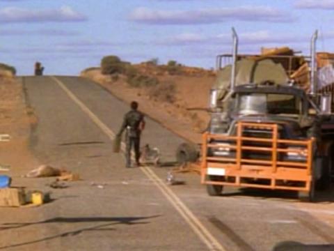 Mad Max stands on the highway next to a truck, holding a tanker of fuel as a motorcycle approaches in the distance