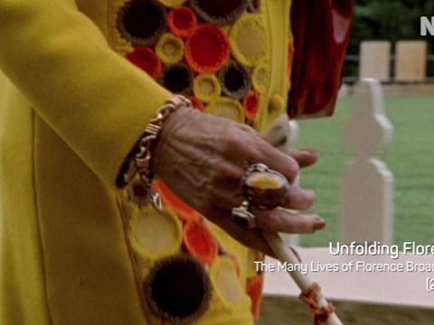 Shot of an elderly woman's arm and hand holding a walking stick. She has a lot of jewellery on and is wearing a brightly coloured knitted top. 