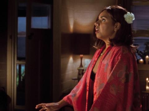 A young Aboriginal woman wearing a red kimono and flower in her hair stands in the middle of a candlelit room facing sideways.
