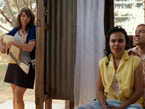 A woman standing in a doorway holding a guitar with two other women are seated to the right of screen. 