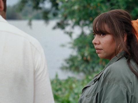 A woman standing by a river with trees behind her turned towards a man standing to her left.