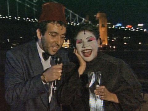 A man and woman standing holding microphones and smiling at the camera. The Sydney Harbour Bridge and skyline is behind them, lit up at night time.