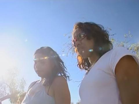 Shot looking up at actor Deborah Mailman and athlete Cathy Freeman who are looking downwards, with a blue sky and trees in the background. 