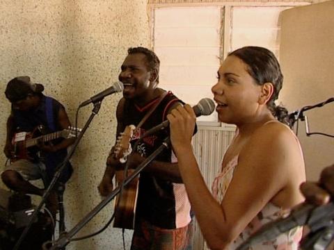 Actor Deborah Mailman at a microphone stand singing with some members of a band.
