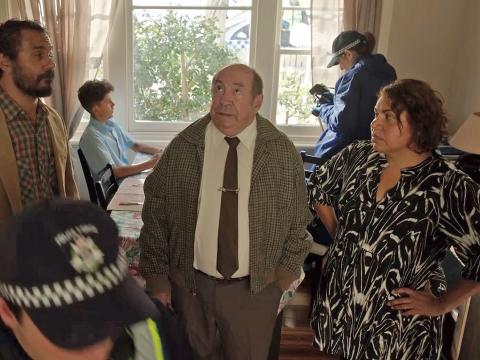 Two police officers look around a room while two men an a woman stand in the centre. A young boy is seated by the window in the background. 