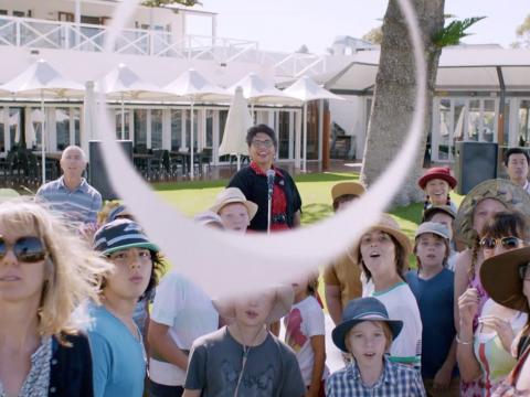 A woman stands in the centre of a group of kids and their parents. She has thrown a circular paper plane which is in the centre of frame.