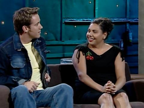 A young man and woman (actors Spencer McLaren and Deborah Mailman) sit on a couch in a TV studio.