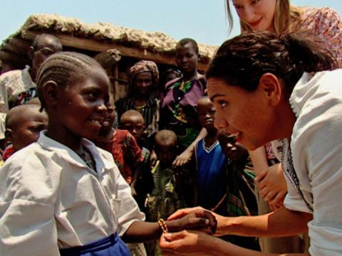 A young African girl receives a gift from actor Deborah Mailman. A group of people from the young girl's community are gathered behind them. 