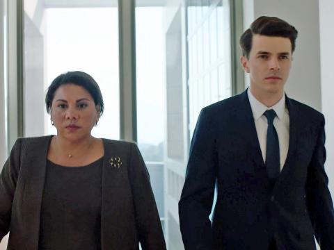 A woman and man walking down a corridor side by side looking serious and determined.