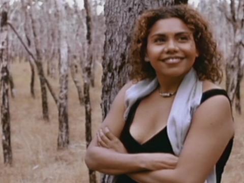 Waist-up shot of actor Deborah Mailman leaning on a tree with her arms folded and smiling.
