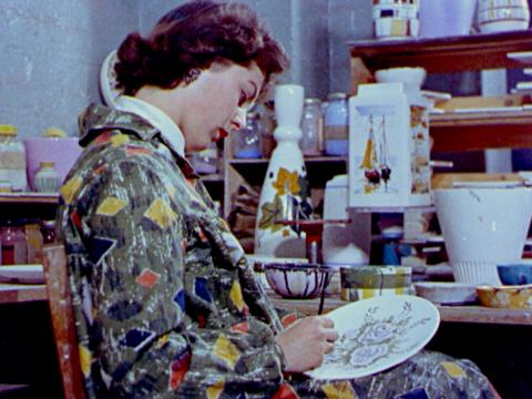 A woman in a colourful smock paints a ceramic plate with a flower design.