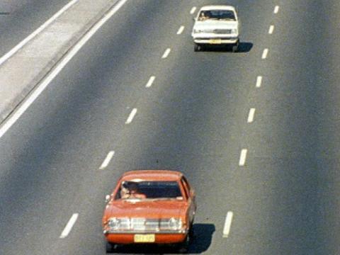Two cars are driving on a three-lane road. A white Torana follows a red Cortina.