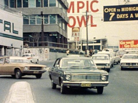 A bronze Holden Kingswood, green and white Falcons, a white Fairlane and other vintage cars on a busy street.