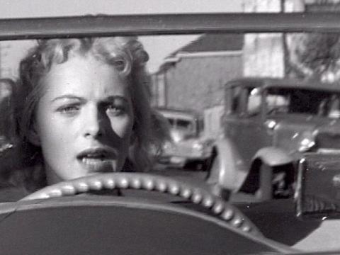 A woman is driving an MG car. She has her mouth open in surprise as a collision is about to take place.