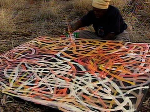Artist Emily Kame Kngwarreye sits outside in the bush doing a painting. Colourful lines representing the pencil yam cover the large canvas which is sitting on the ground in front of her.