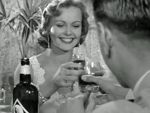 A woman smiles as she charges her glass and does a 'cheers' with a man (seen from behind). They are in a club with a plant in the background.