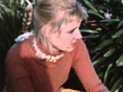 A blonde-haired woman wearing an orange jumper sits in a garden.