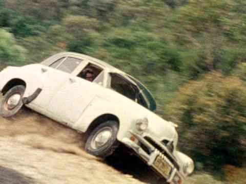 A vintage FJ Holden car is rolling off the road towards the bush below in a car accident.